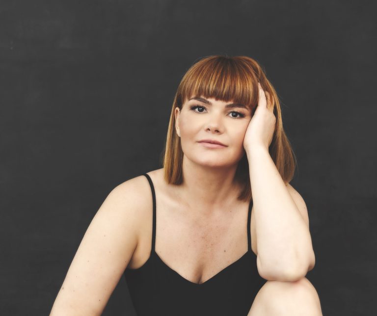 Portret feminin al unei femei roșcate, într-o ținută neagră, cu mâna sprijinită de cap și privirea îndreptată spre cameră.